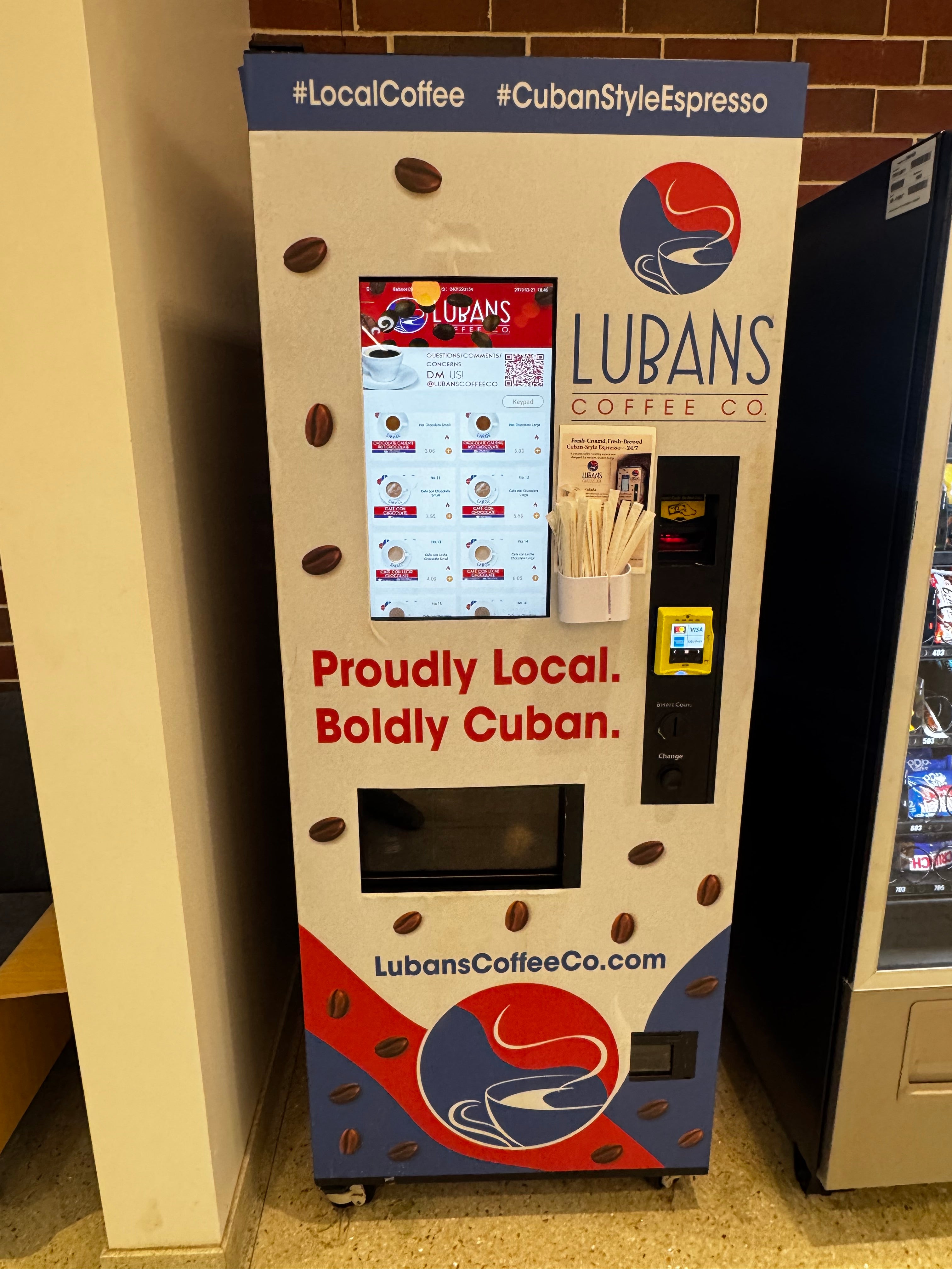 A Lubans Coffee Co. espresso coffee vending machine photographed in a real indoor environment, shown sideways with surrounding floor and counter visible.